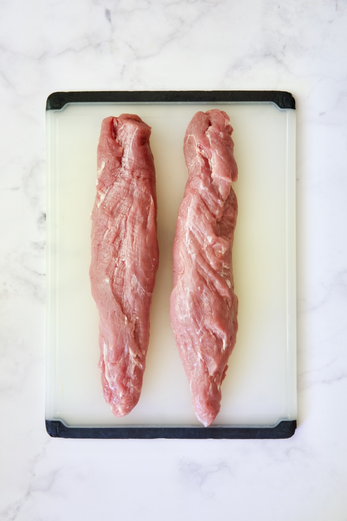 Two trimmed pork tenderloins on white plastic cutting board.