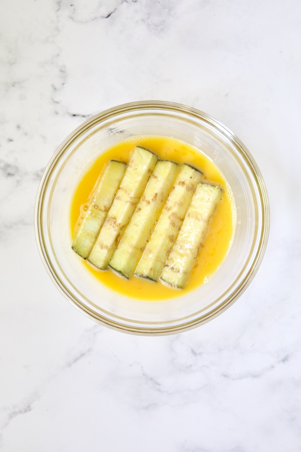 Eggplant fries in beaten egg in glass bowl.