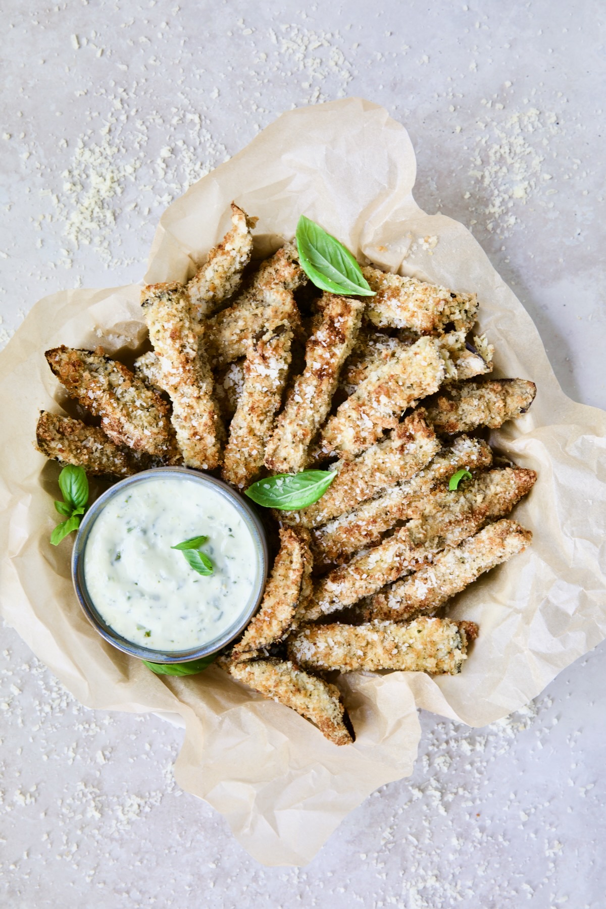 Air Fryer Eggplant Fries with Garlic Basil Aioli in parchment paper lined basket with Parmesan cheese scattered about.