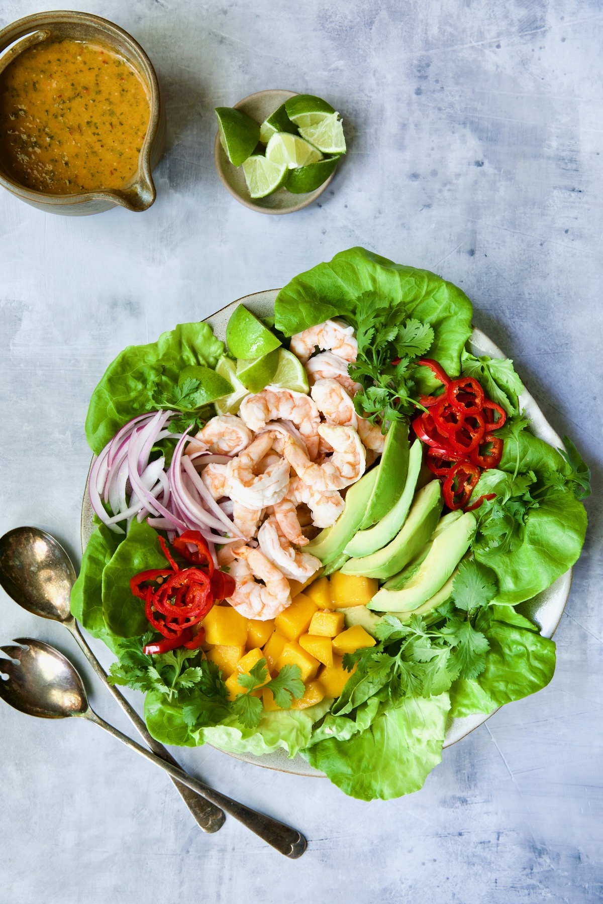 Butter Lettuce Salad with Shrimp, Mango and Avocado on blue background with vinaigrette and lime wedges.