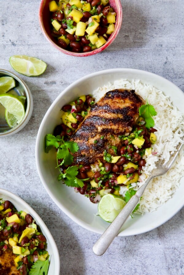 Caribbean Jerk Chicken with Mango Black Bean Salsa in white bowl garnished with cilantro and squeezed lime wedges.
