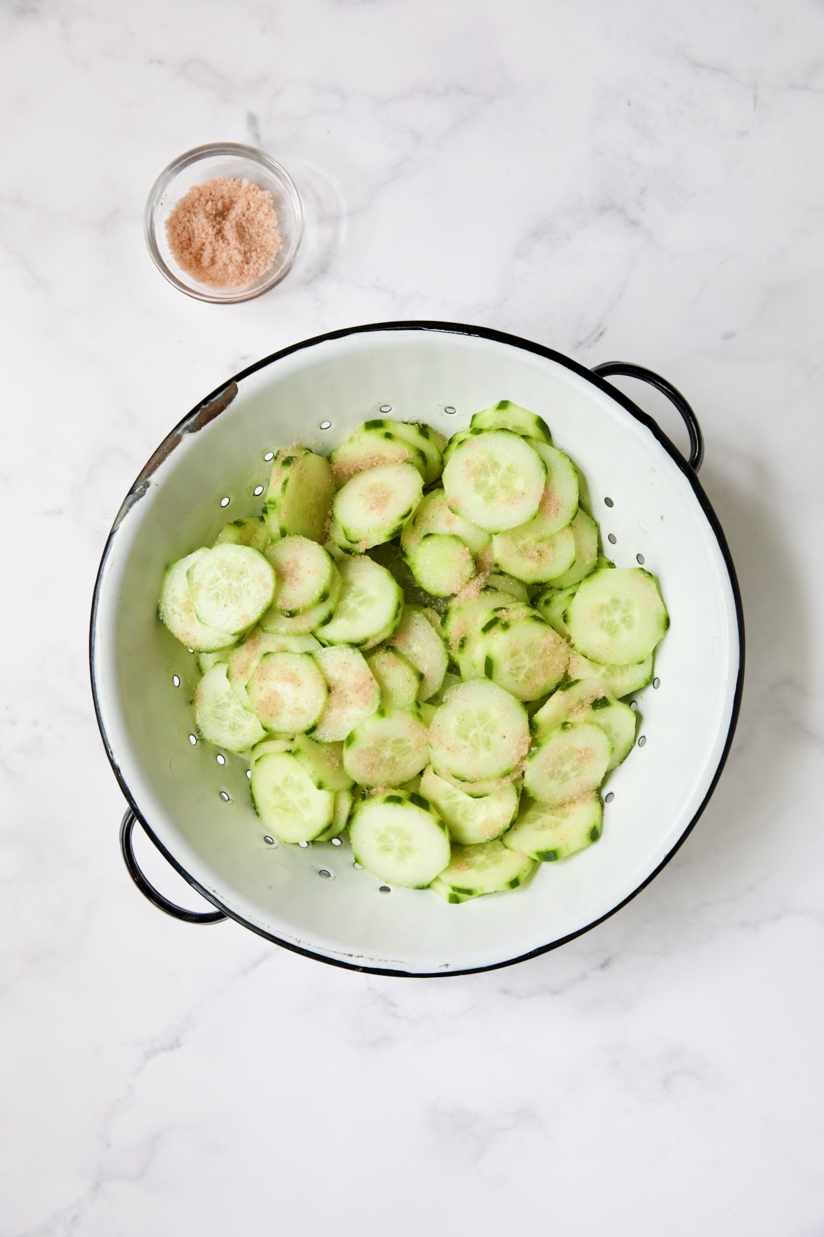 Cucumber in white distressed colander with salt sprinkled over them.