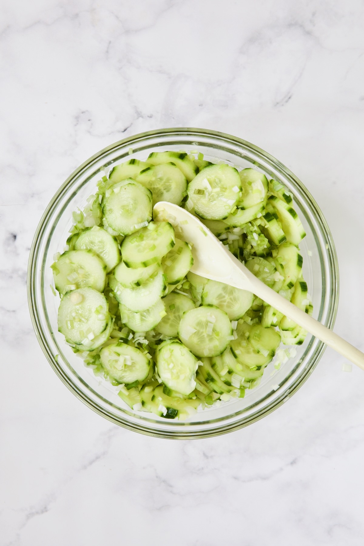 Cucumbers and scallions being stirred with white spoon in glass bowl.