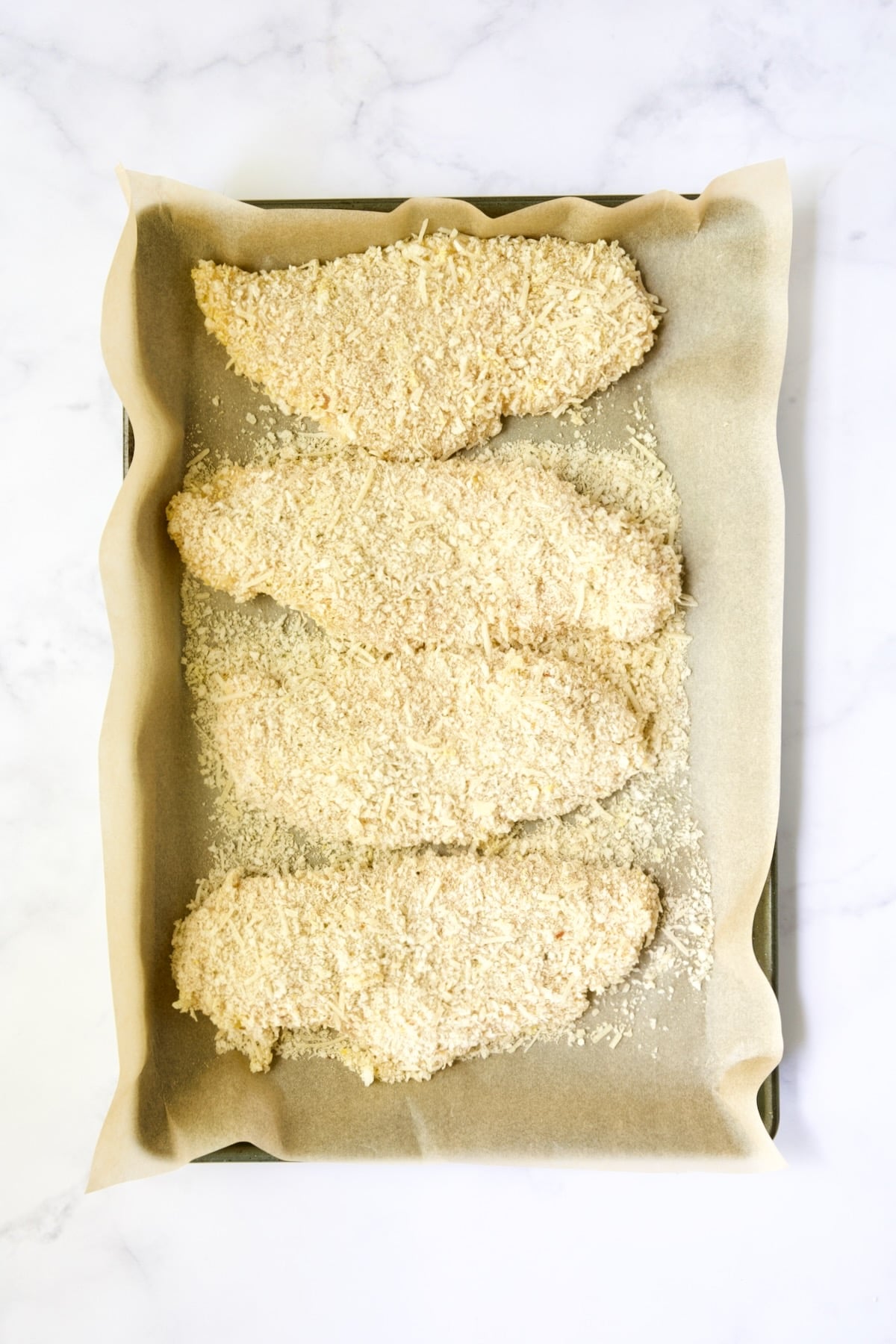 Four prepped pieces of breaded chicken on parchment paper-lined baking sheet.