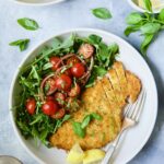 Chicken Milanese with Arugula and Cherry Tomato Salad garnished with fresh basil sprigs and lemon wedges.