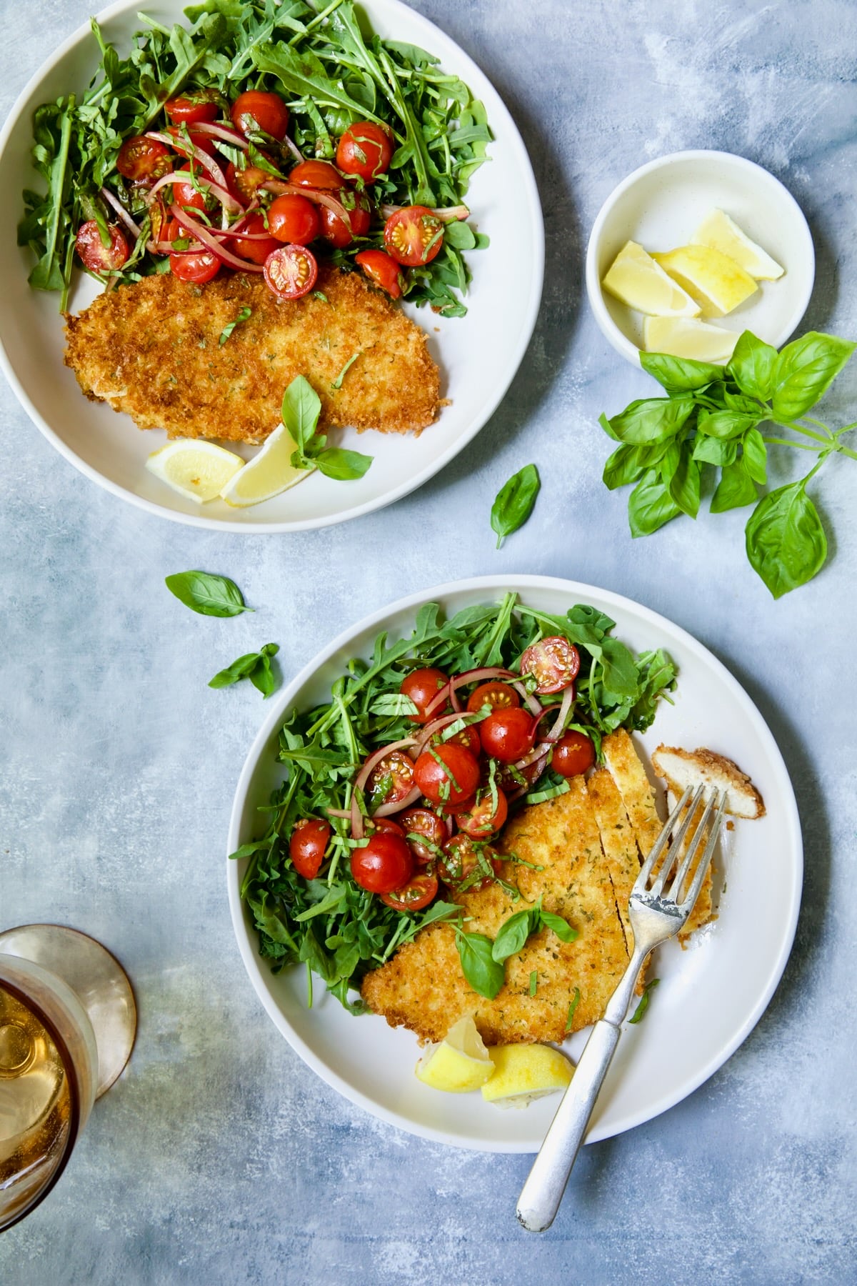 Two servings of Chicken Milanese with Arugula and Cherry Tomato Salad on white plates garnished with fresh basil and lemon wedges.