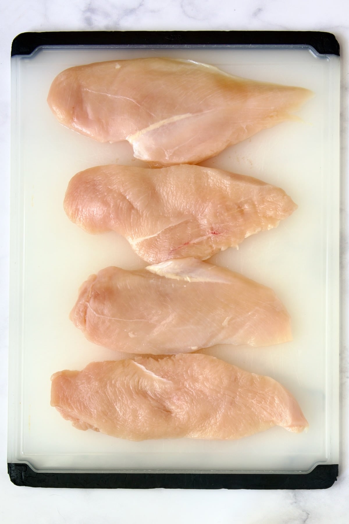 Four chicken cutlets on white plastic cutting board.