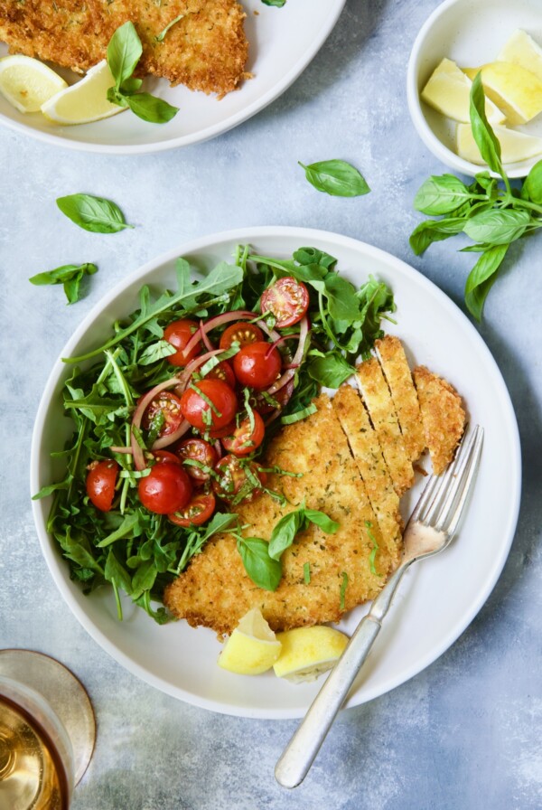 Chicken Milanese with Arugula and Cherry Tomato Salad garnished with fresh basil sprigs and lemon wedges.