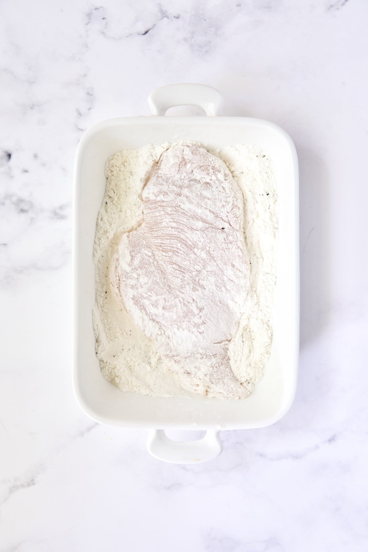 Chicken cutlet coated with flour in white dish.