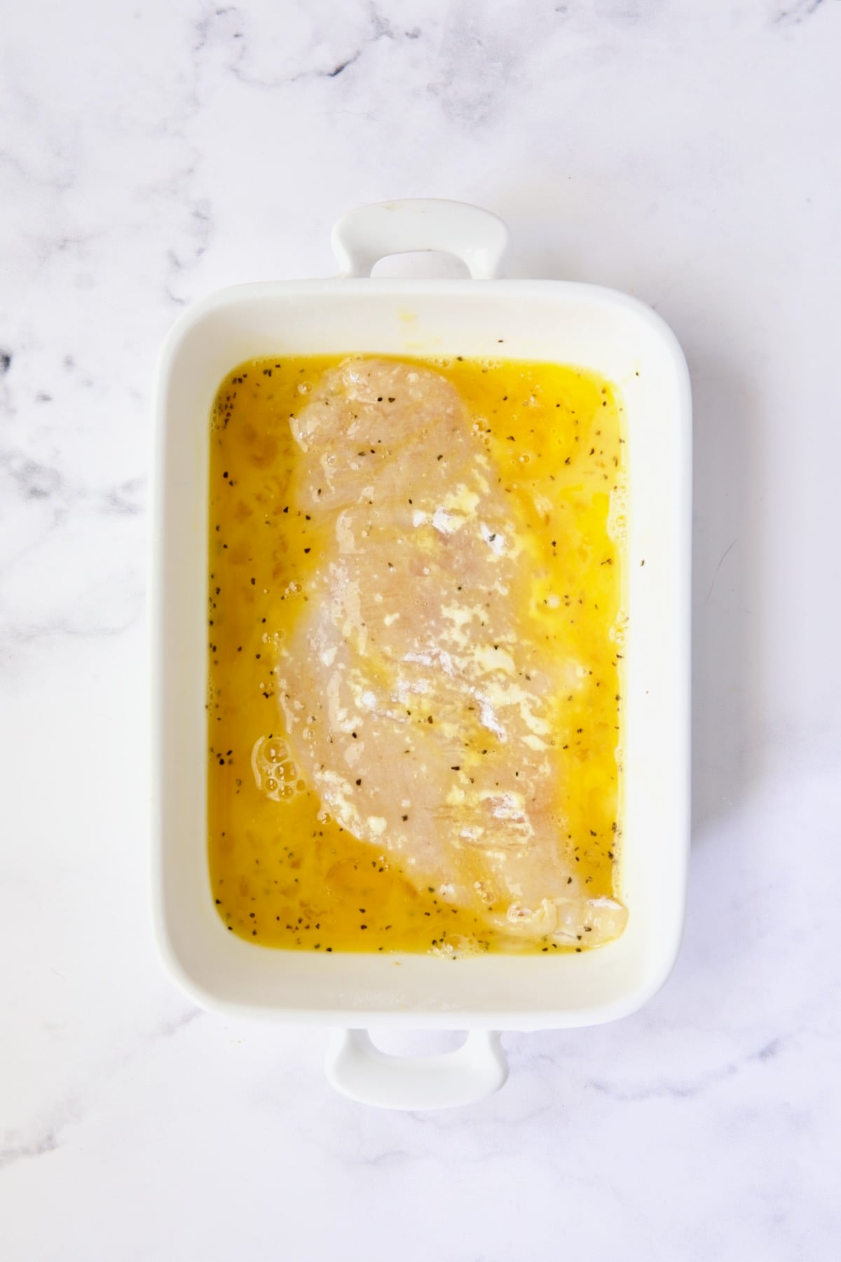 Chicken cutlet in egg mixture in white dish.