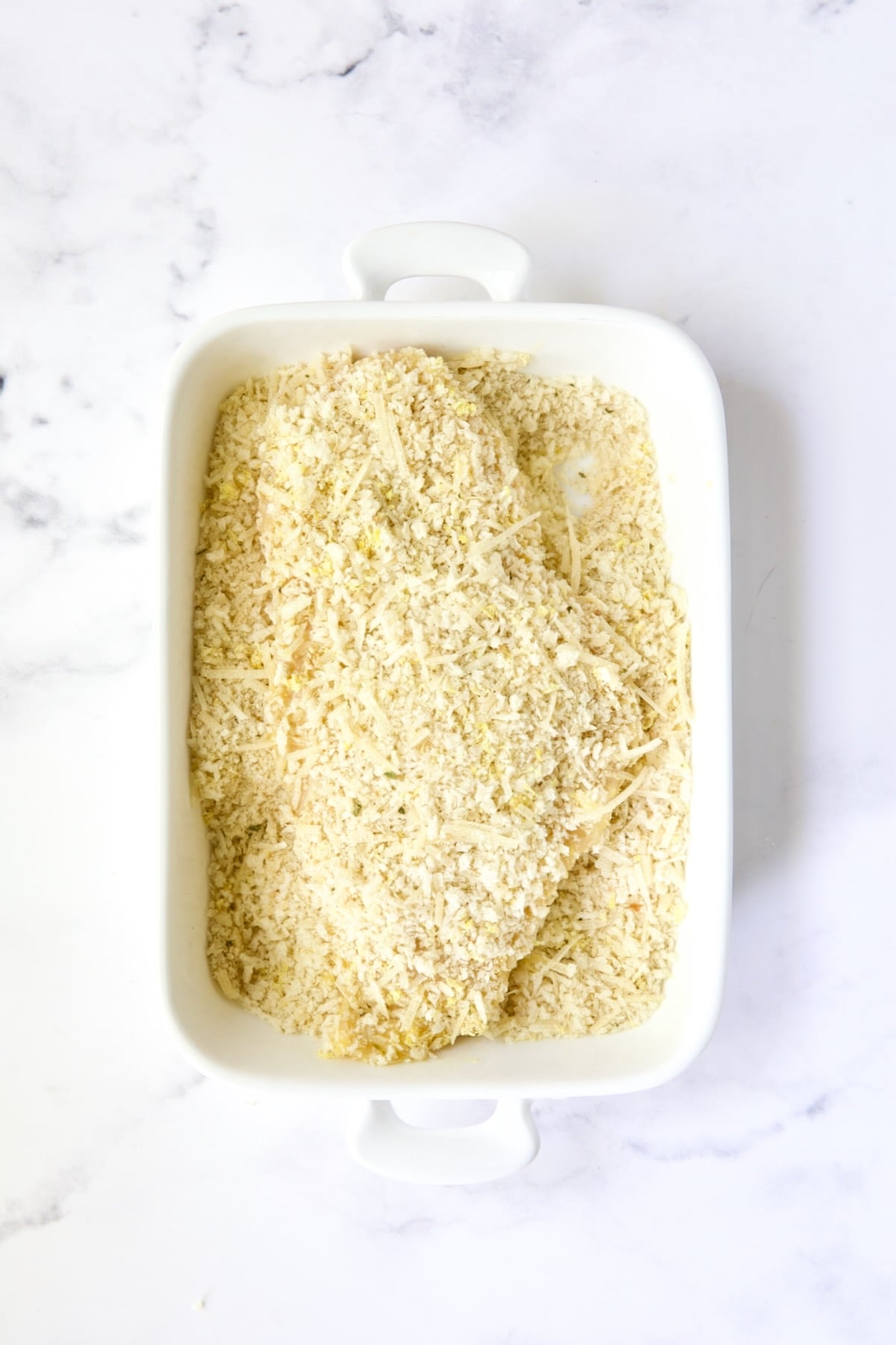 Uncooked breaded chicken cutlet in white rectangular dish in crumbs..