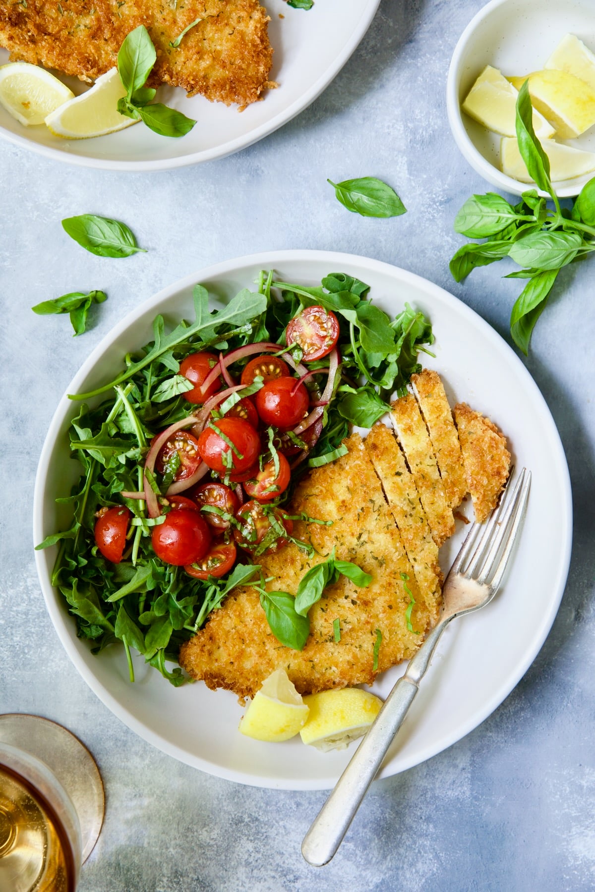 Chicken Milanese with Arugula and Cherry Tomato Salad garnished with fresh basil sprigs and lemon wedges.