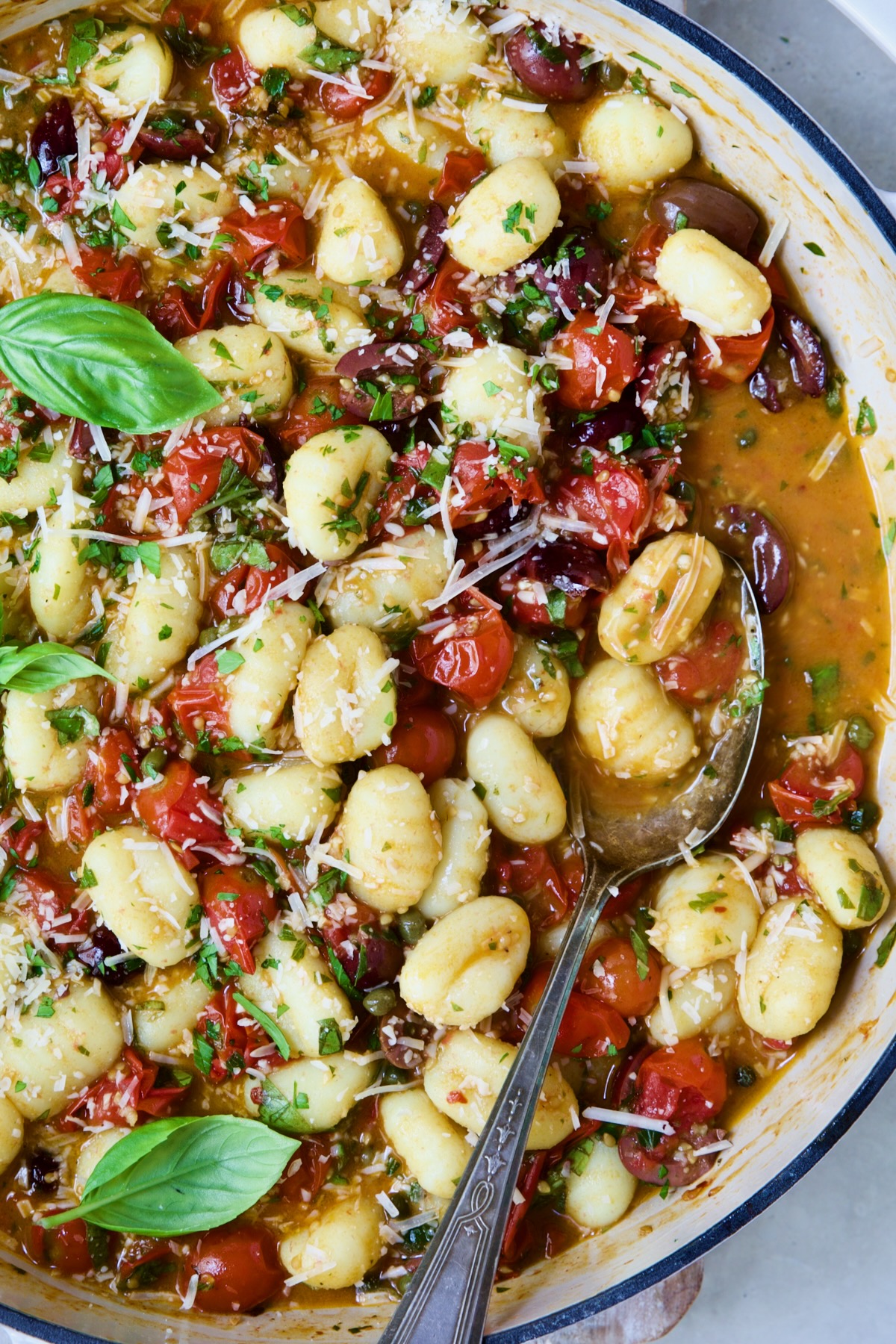 Gnocchi with Cherry Tomato Puttanesca Sauce with some on serving spoon in white enamel cast iron skillet.