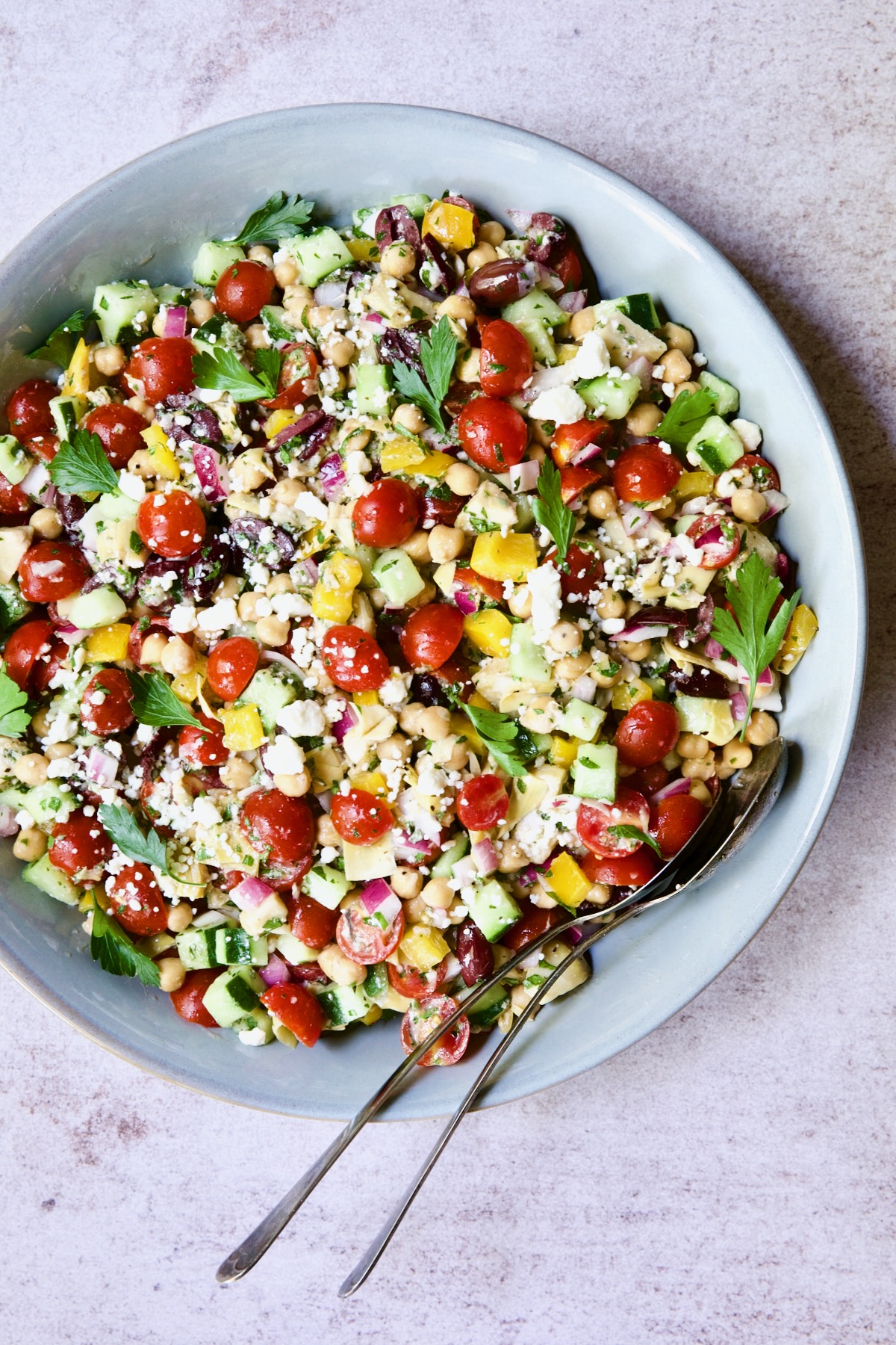 Mediterranean Chopped Salad with Chickpeas in blue bowl with serving utensils garnished with fresh parsley leaves.
