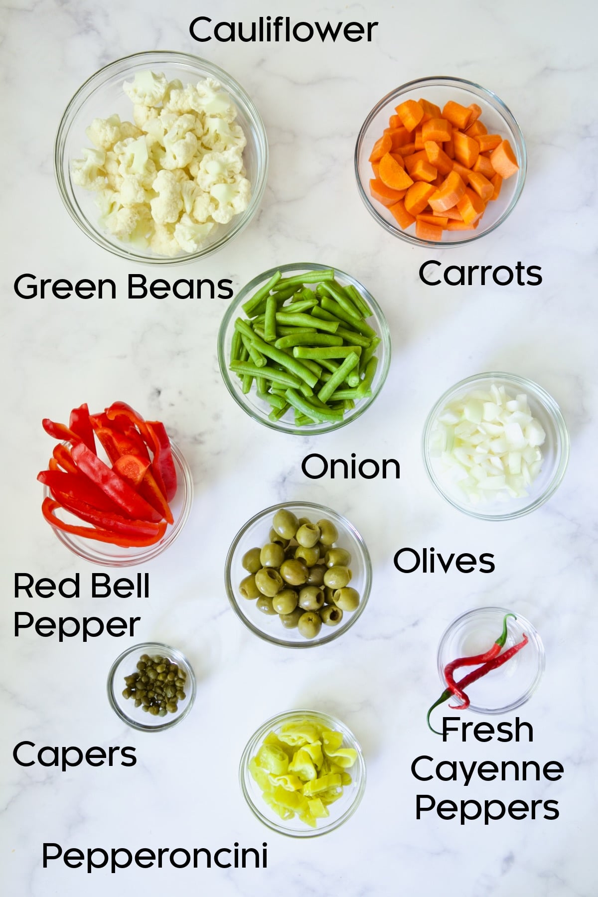 Cauliflower, carrots, green beans, red bell pepper, onion, olives, capers, pepperoncini and cayenne peppers in glass bowls.