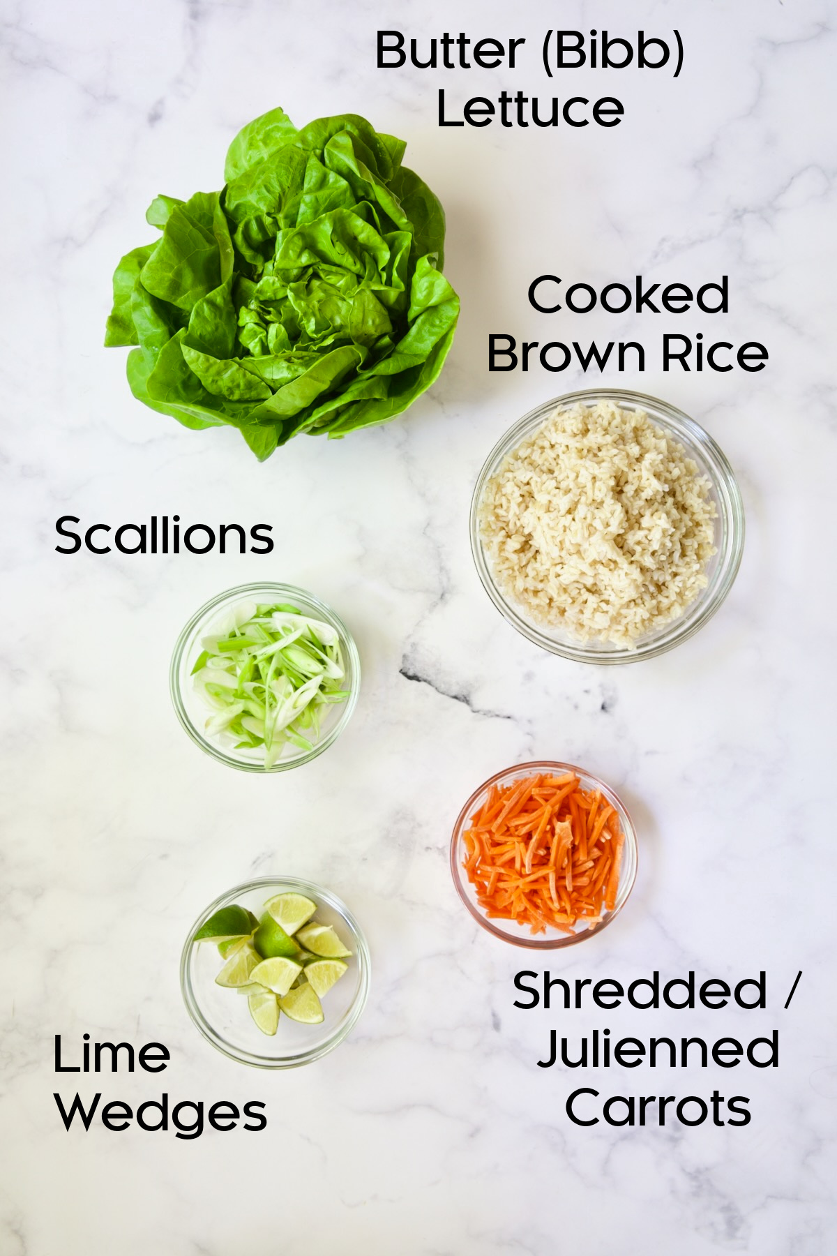 Lettuce, cooked brown rice, scallions, lime wedges and julienned carrots in glass bowls.