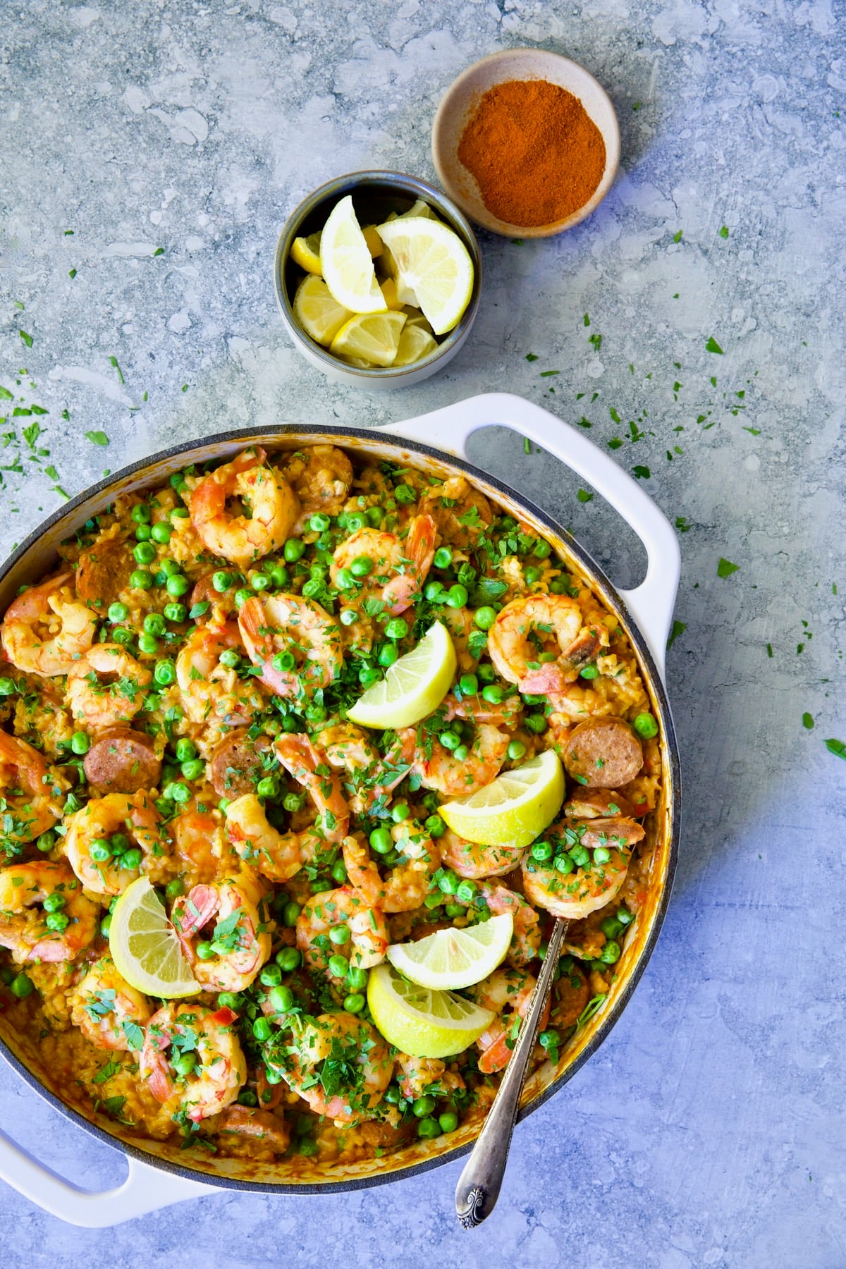 Skillet of Brown Rice Paella with Shrimp and Chorizo with lemon wedges and bowl of paprika.