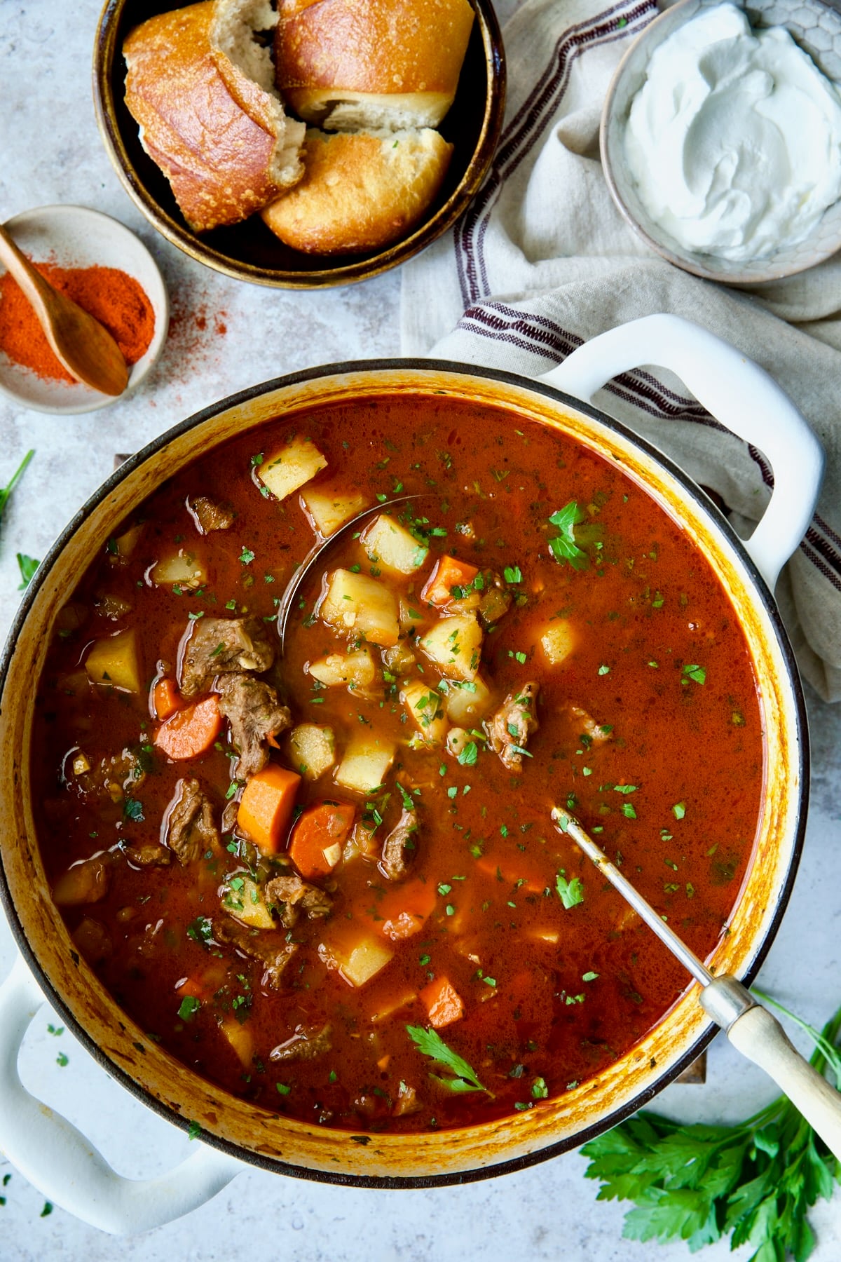 Finished Traditional Hungarian Goulash Soup Recipe in white Dutch oven with bread and sour cream.
