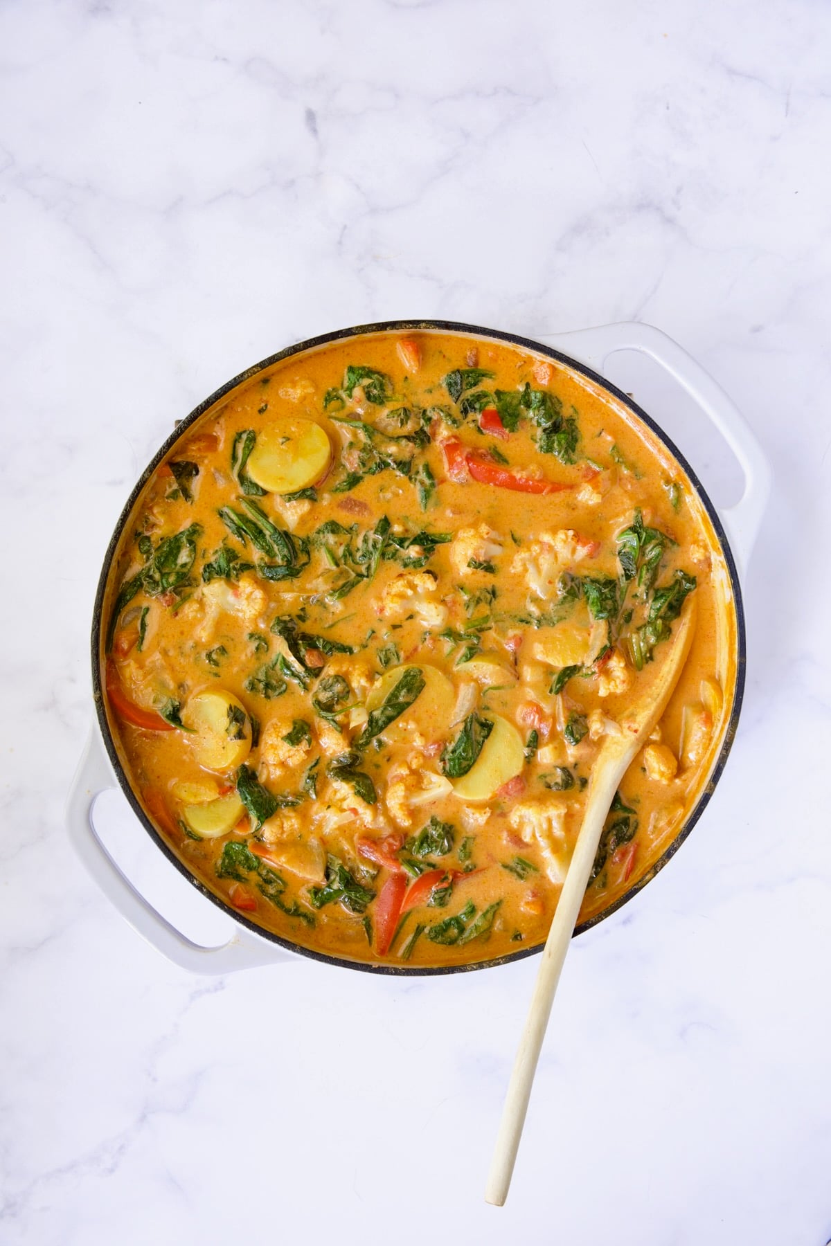 Vegetable curry in white enamel cast iron skillet being stirred with white spoon.
