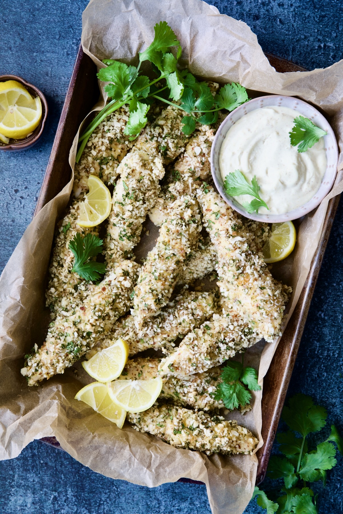Almond-Crusted Chicken Tenders with Lemon Curry Mayonnaise in parchment paper lined baking pan with cilantro and lemon wedges.