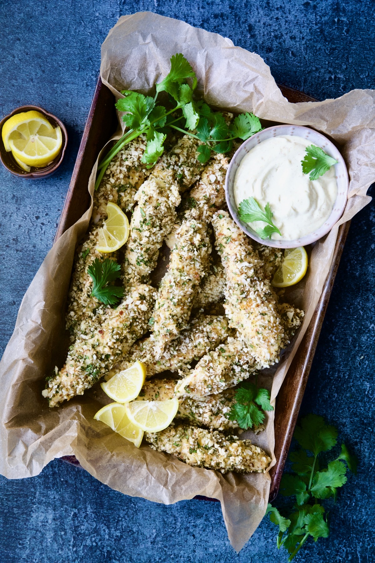 Almond-Crusted Chicken Tenders with Lemon Curry Mayonnaise in parchment paper-lined pan garnished with lemon wedges and cilantro.