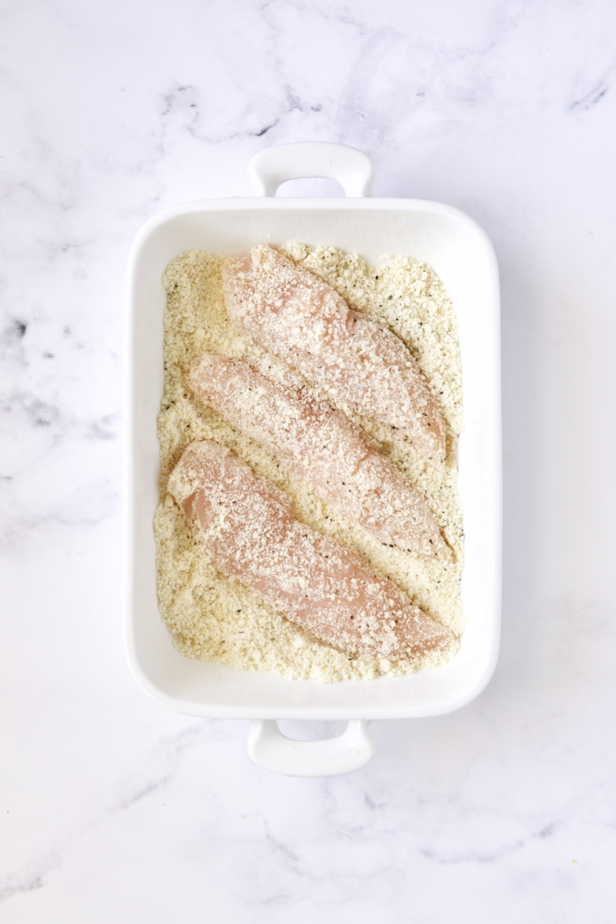 Three chicken tenders in almond flour in white rectangular baking pan.