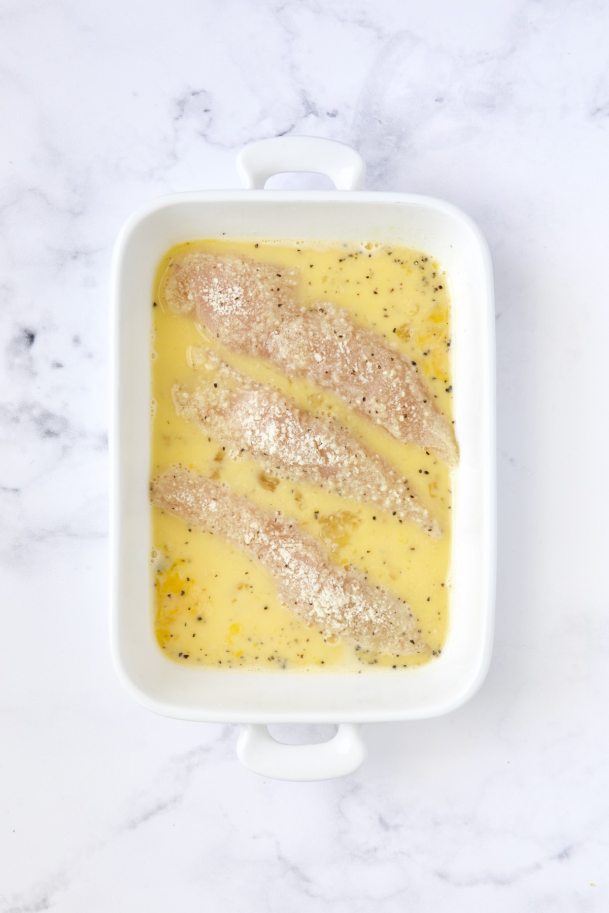 Three chicken tenders in beaten egg after being coated with almond flour in white rectangular baking pan.