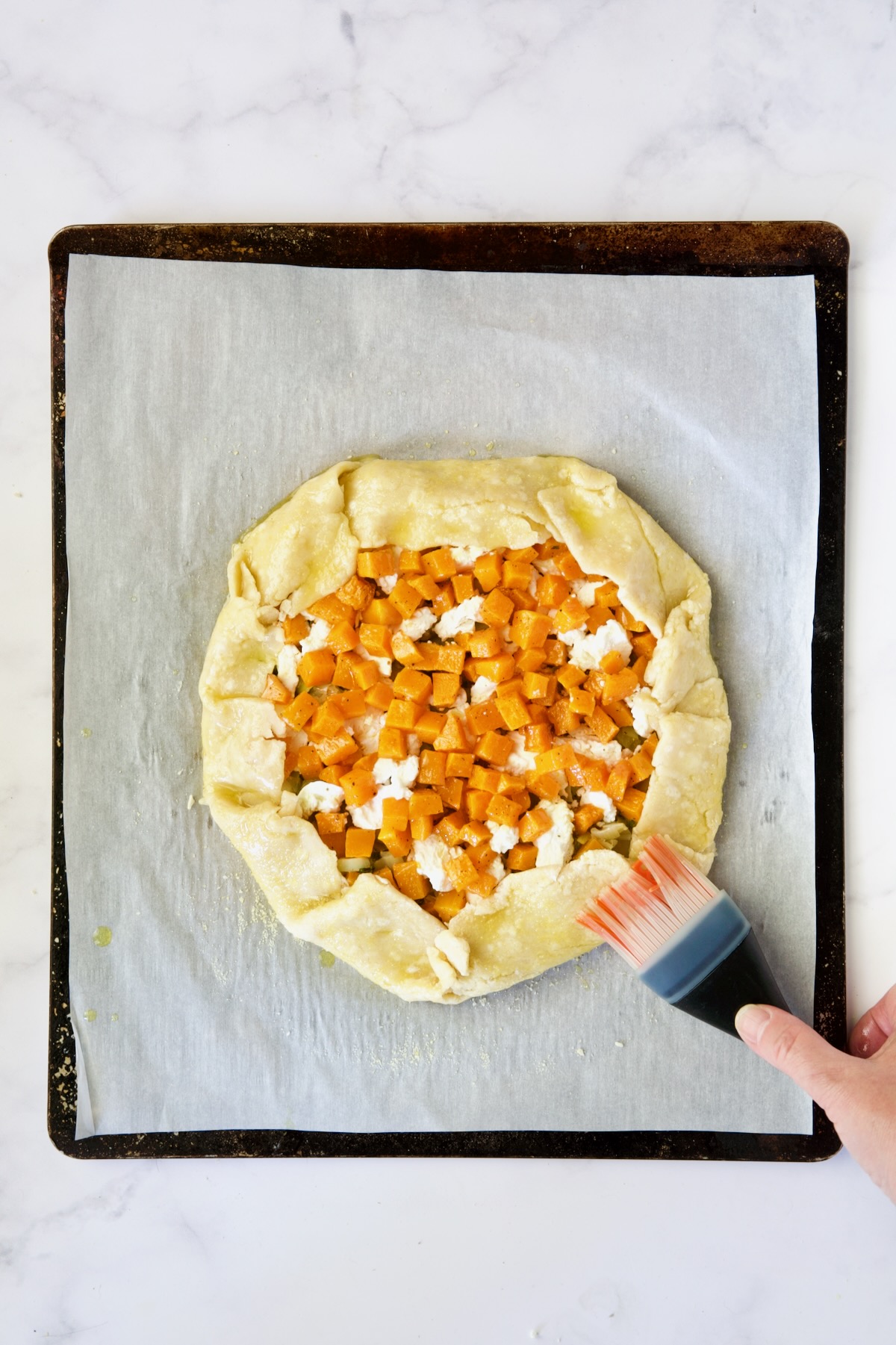 Assembled galette on white parchment paper-lined baking sheet being brushed with olive oil.