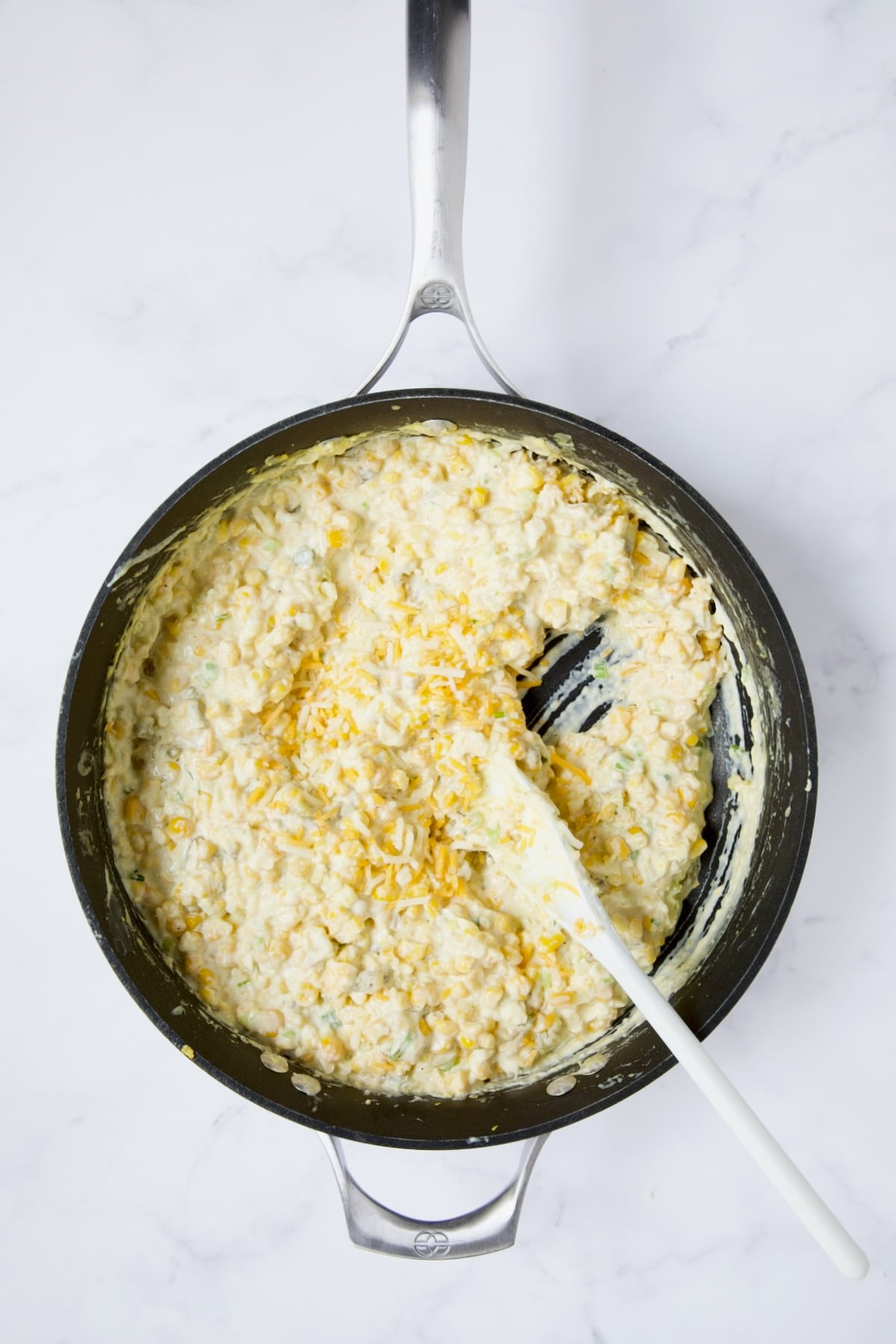 Corn casserole in nonstick skillet with some of the cheese added being stirred with white spoon.