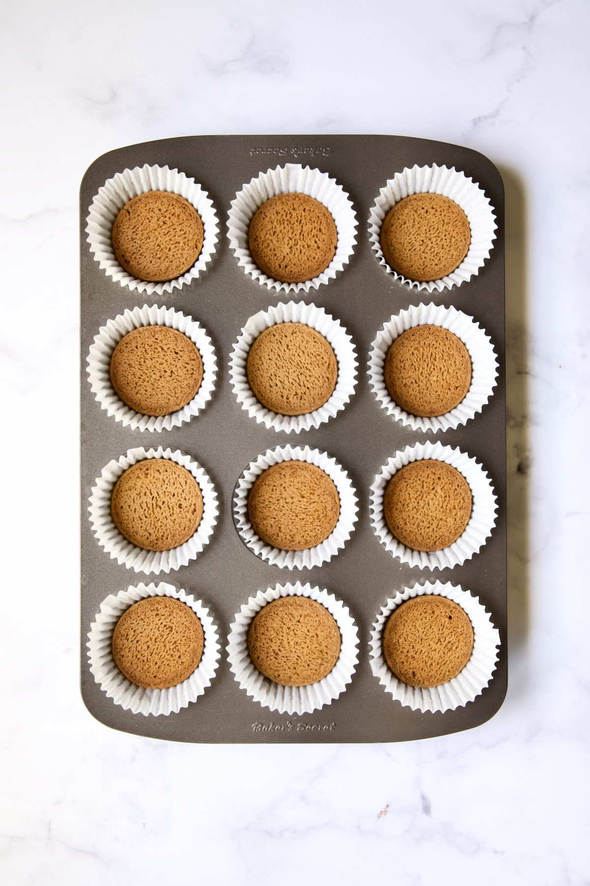 Muffin pan lined with white paper cups with a gingersnap cookie placed in the bottom of each cup.