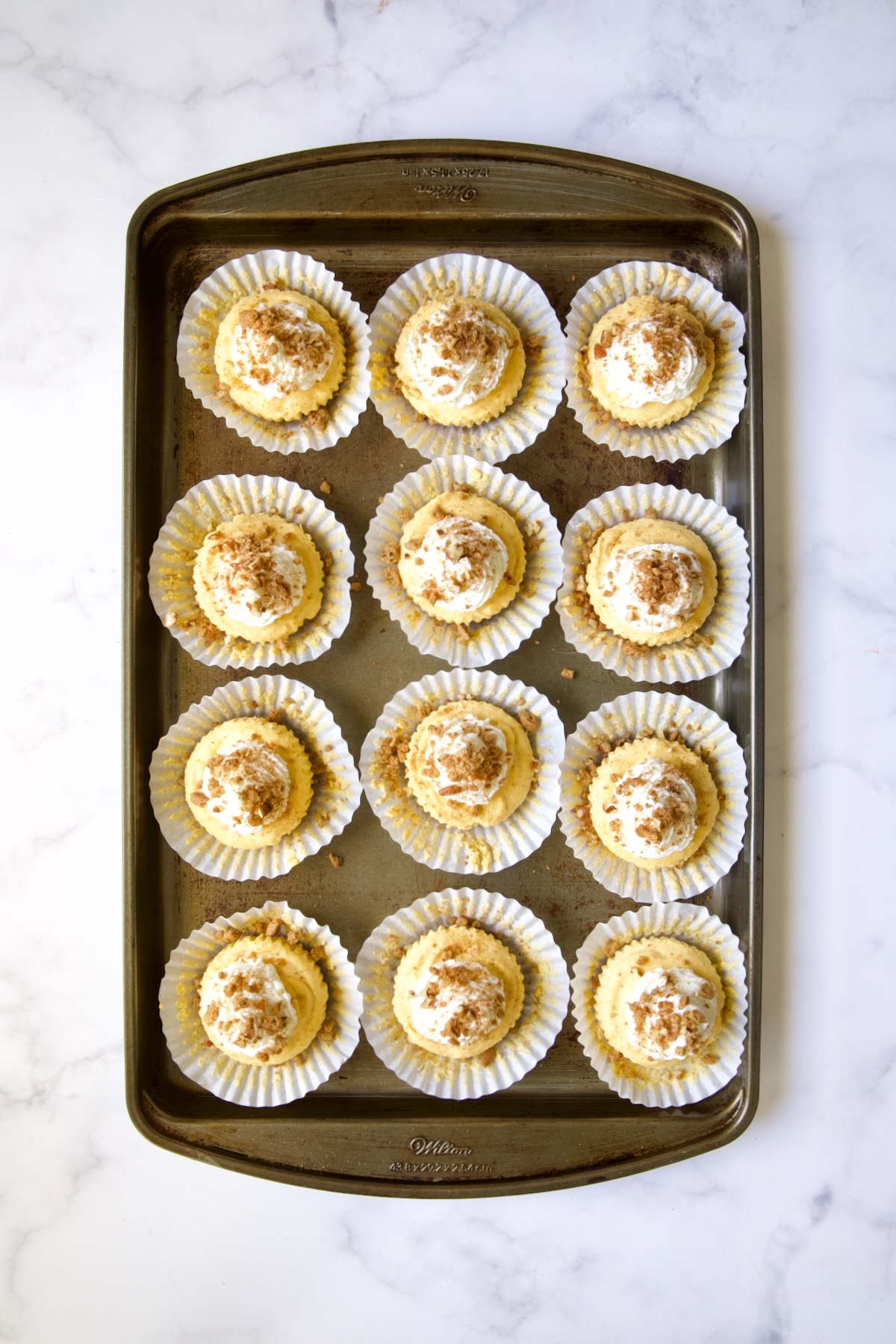 Twelve set cheesecakes on white muffin pan liners on baking sheet with topping sprinkled over.