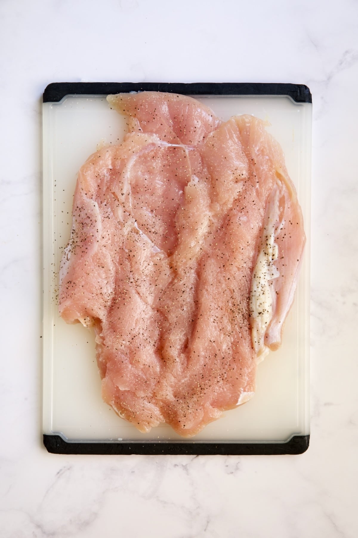 Butterflied turkey breast seasoned with salt and pepper on white plastic cutting board.