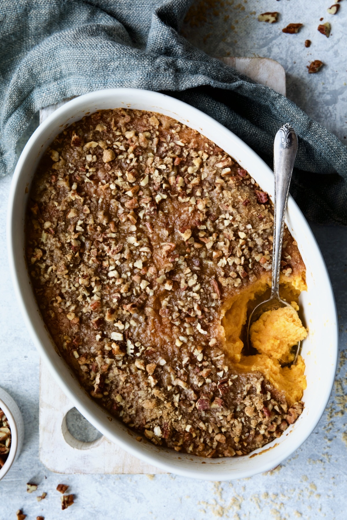 Sweet Potato Souffle in oval baking dish with blue napkin and pecans and brown sugar scattered about.