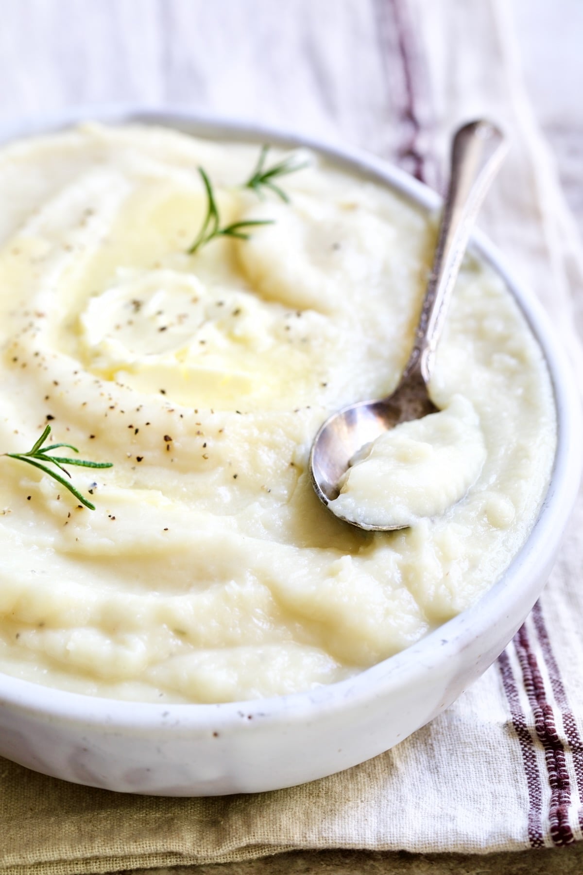 Creamy Mashed Turnip Recipe in white oval bowl with serving spoon on napkin with red stripe garnished with fresh rosemary.