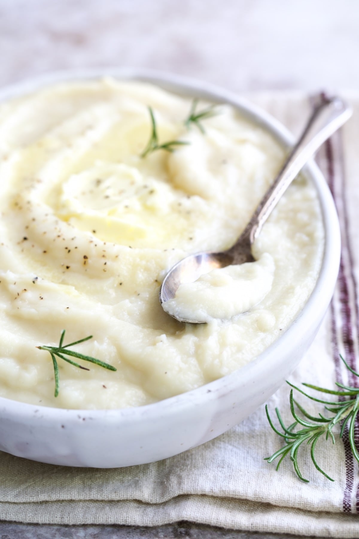 Creamy Mashed Turnip Recipe in white oval bowl with serving spoon on napkin with red stripe garnished with fresh rosemary.