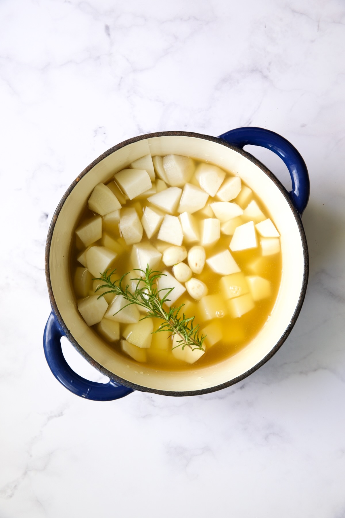 Cubed turnips, potatoes and a sprig of rosemary in blue Dutch oven with chicken broth.