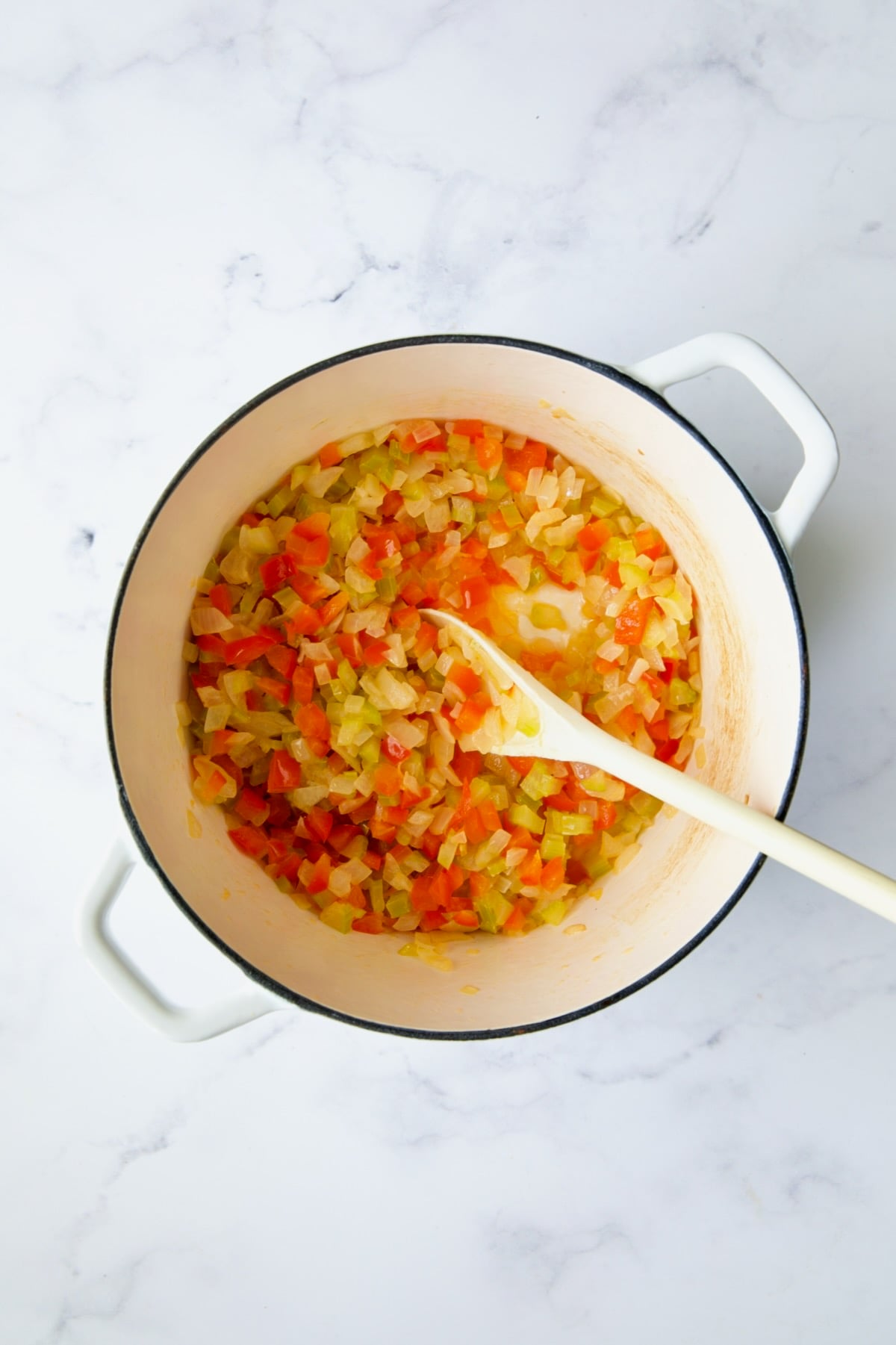 Cooked onions, celery and red bell pepper in white Dutch oven being stirred with white spoon.