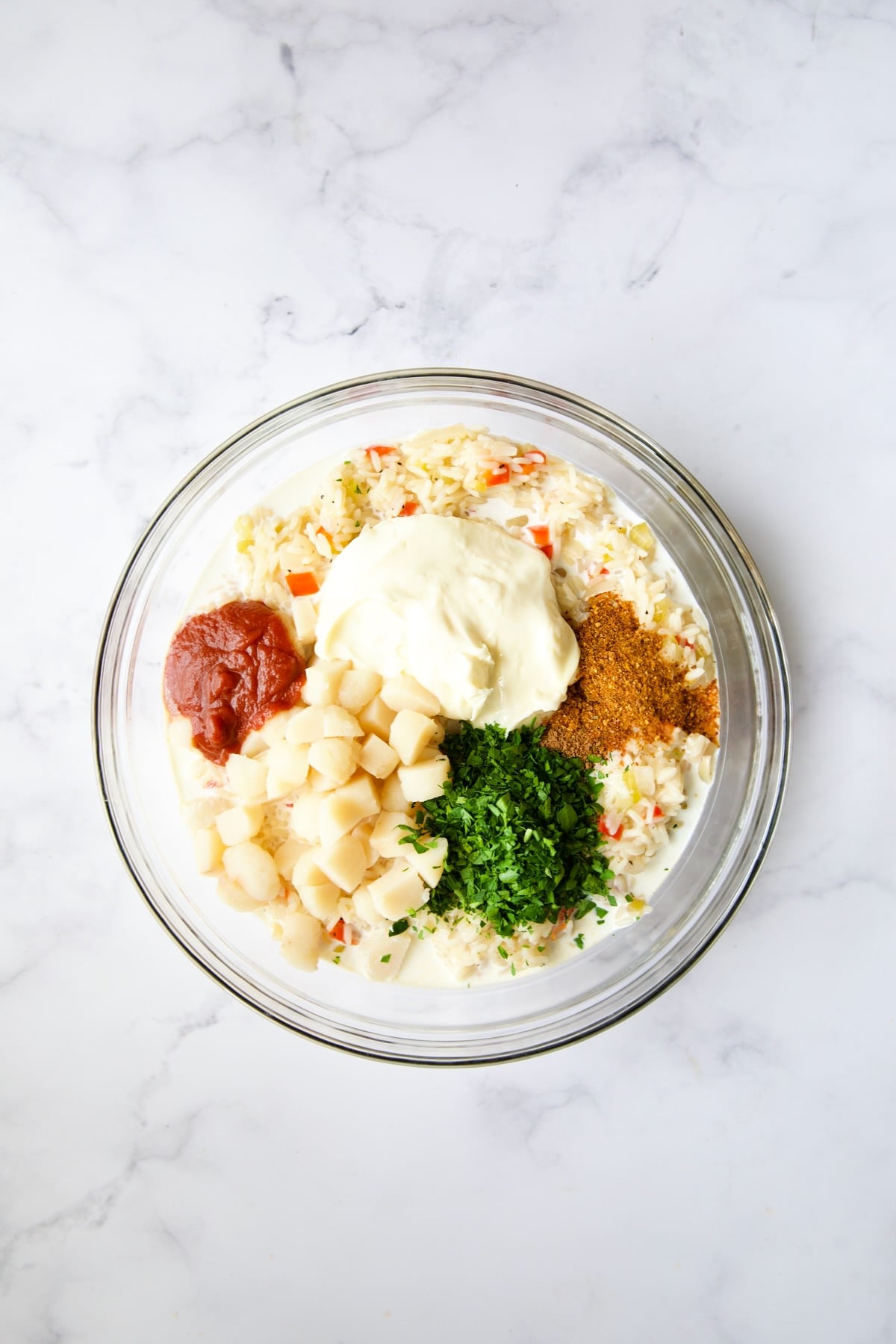 Cooked rice and vegetables in glass bowl with half-and-half, mayonnaise, tomato puree, parsley, water chestnuts and spices in glass bowl.