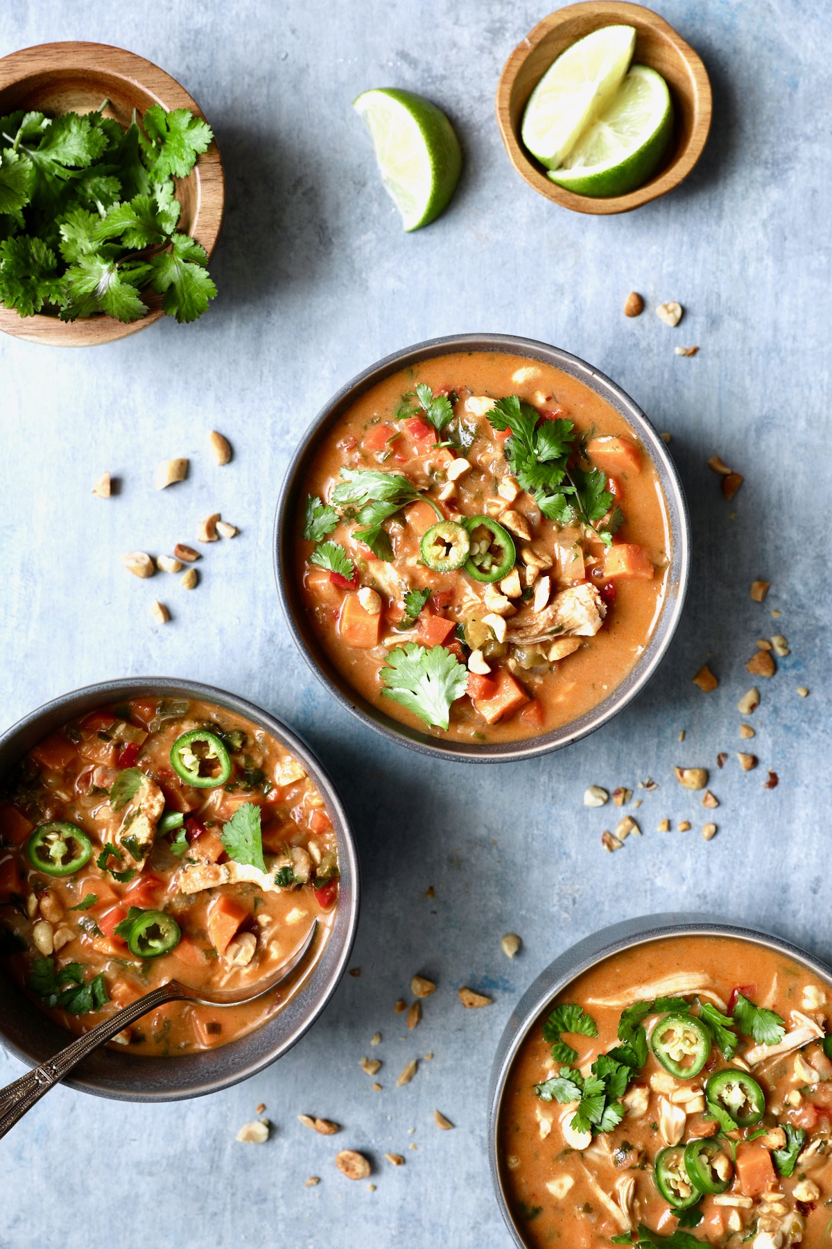 Three bowls of Spicy Chicken, Sweet Potato and Peanut Soup in gray bowls on blue background.