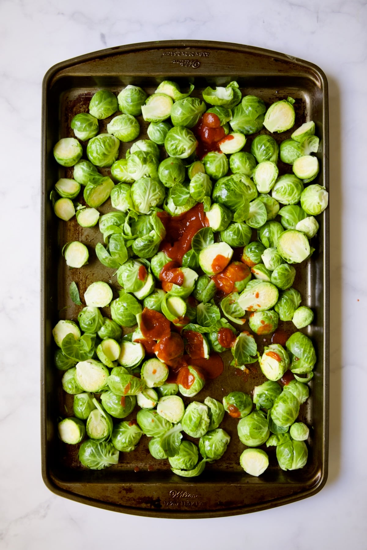 Halved Brussels sprouts on baking sheet before being roasted with Sriracha poured over them.