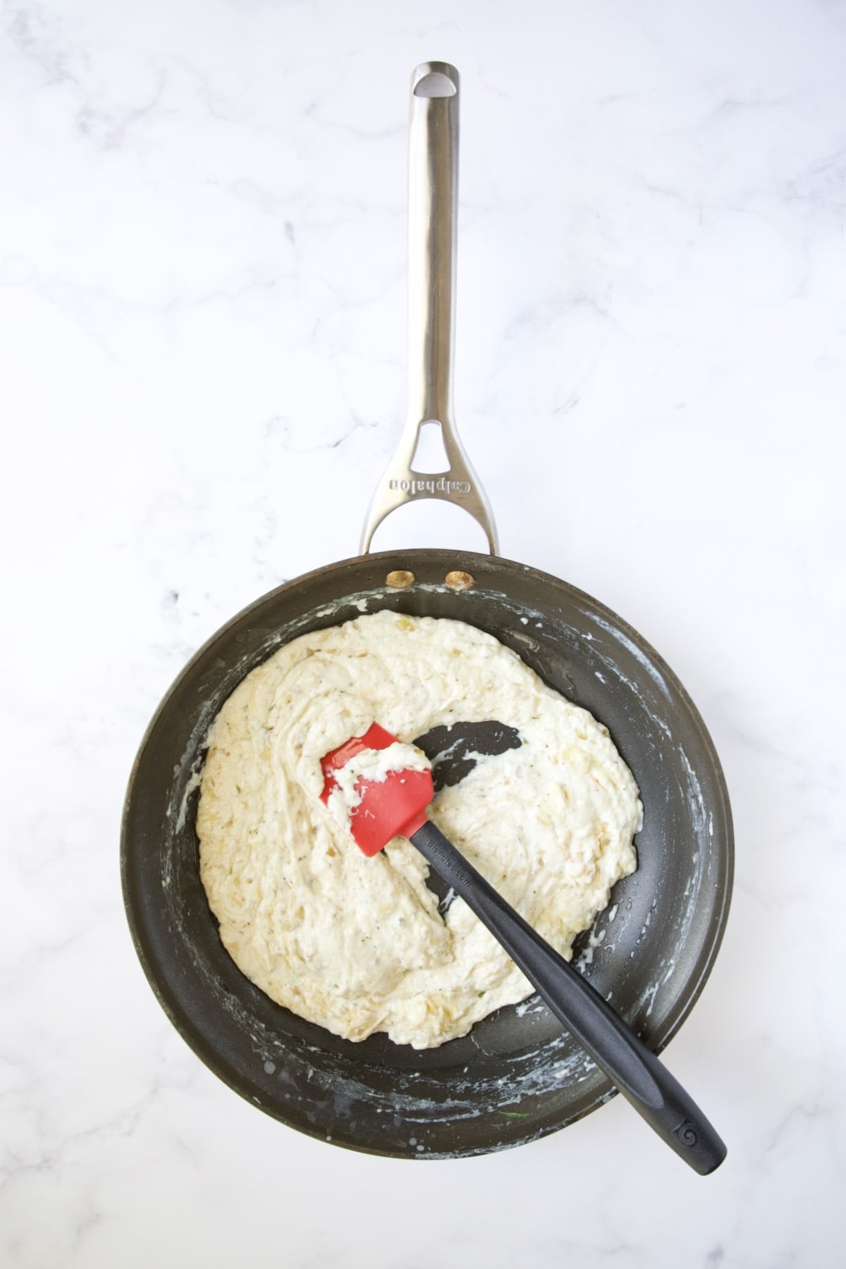 Thickened cheese sauce in nonstick skillet being stirred with red and black spatula.