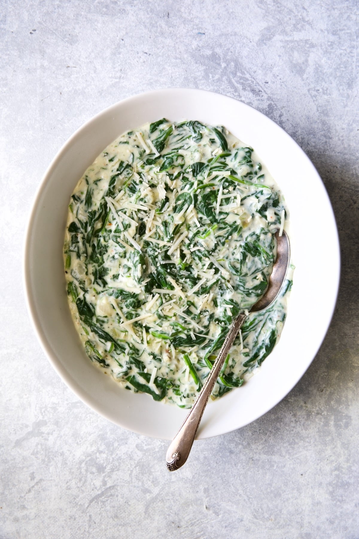 Steakhouse Creamed Spinach in white oval bowl with serving spoon on light blue-gray background.