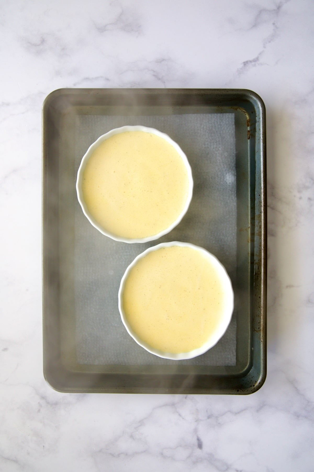 Two creme brulee dishes with boiling water added to rimmed sheet pan before being baked.