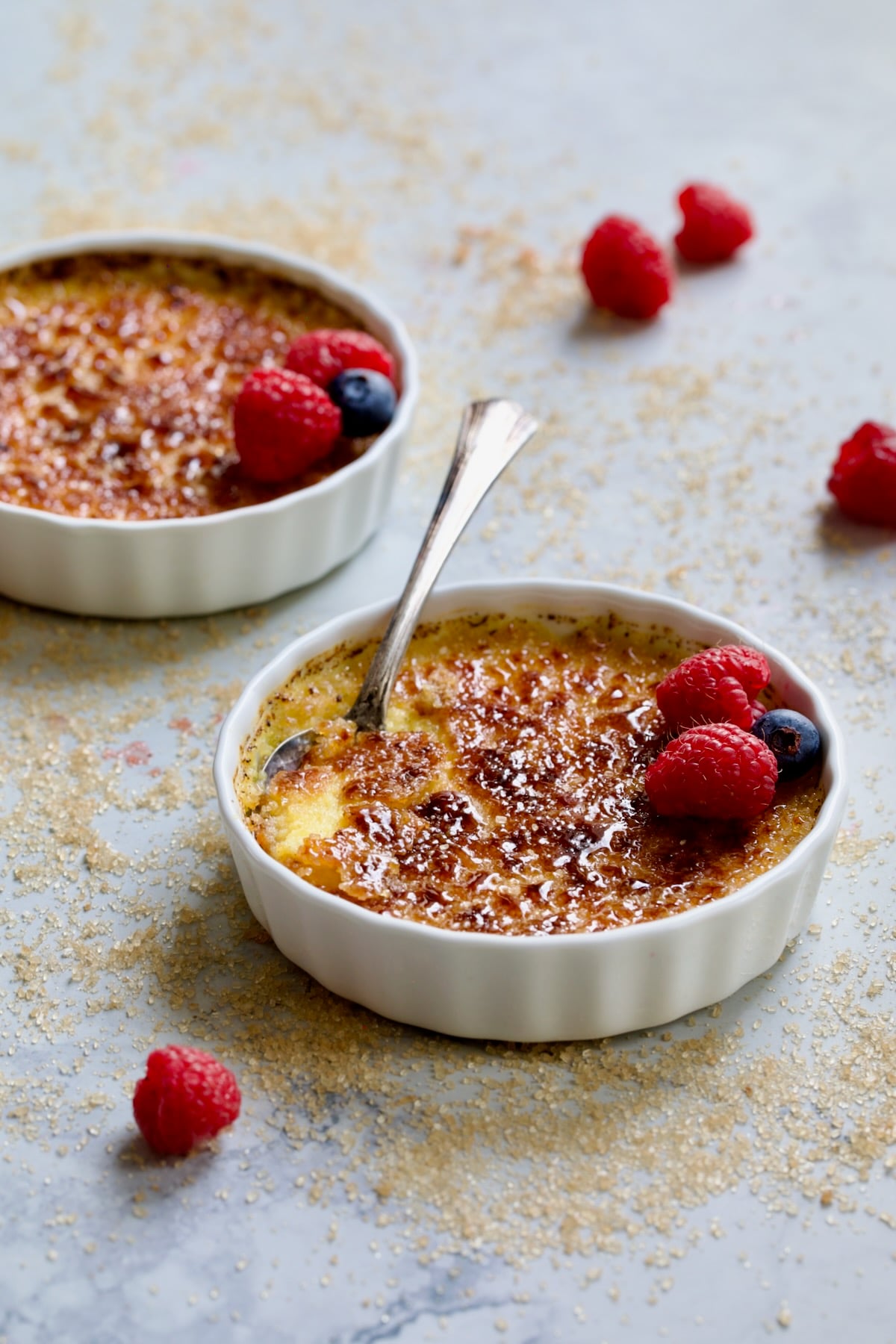Two dishes of Classic Creme Brulee for Two garnished with red raspberries and blueberries with sugar scattered over the surface.