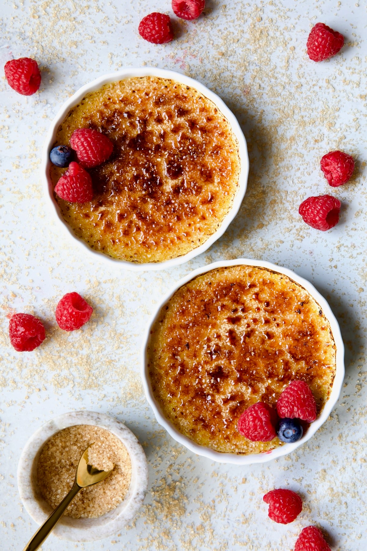 Two dishes of Classic Creme Brulee for Two garnished with red raspberries and blueberries with small bowl of sugar.