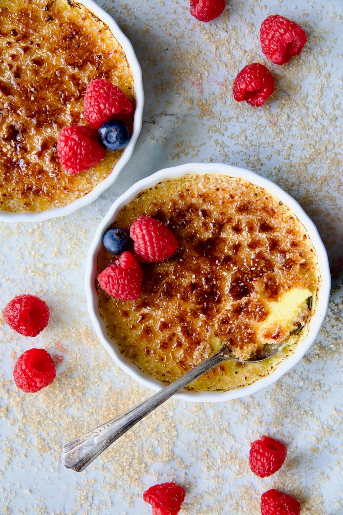 Two dishes of Classic Creme Brulee for Two garnished with red raspberries and blueberries with spoon breaking into one of the dishes.