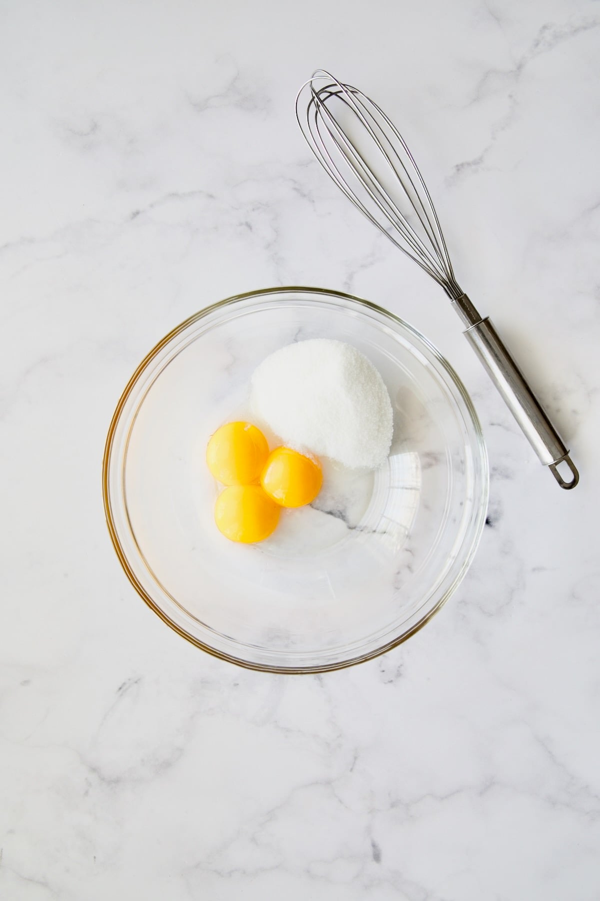 Three egg yolks and granulated sugar in glass bowl with wire whisk.