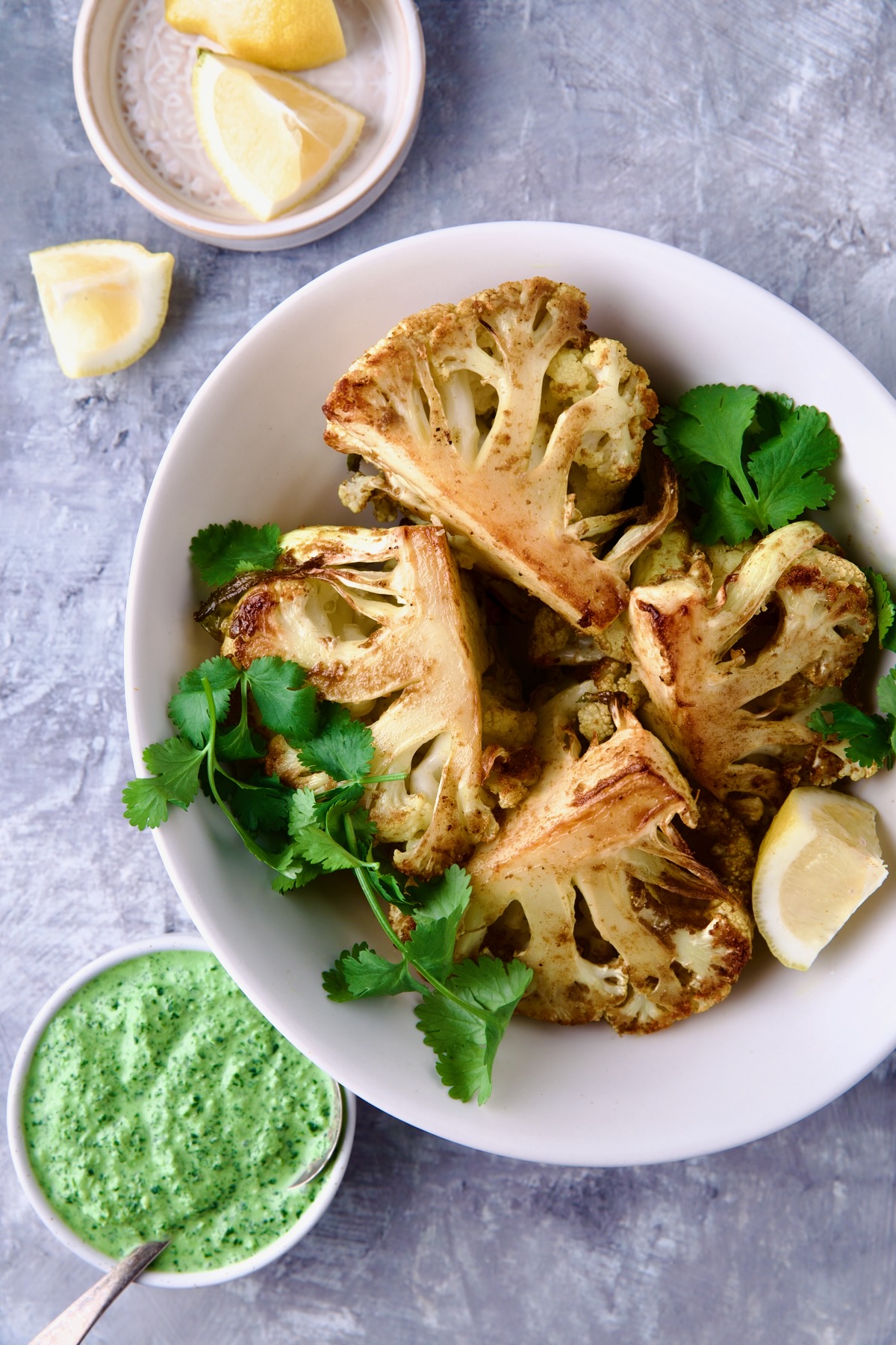 Four wedges of Curry Roasted Cauliflower in white oval bowl with Cilantro Chutney and lemon wedges.