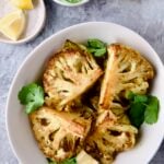 Four wedges of Curry Roasted Cauliflower Wedges with Cilantro Chutney in white bowl with lemon wedges.