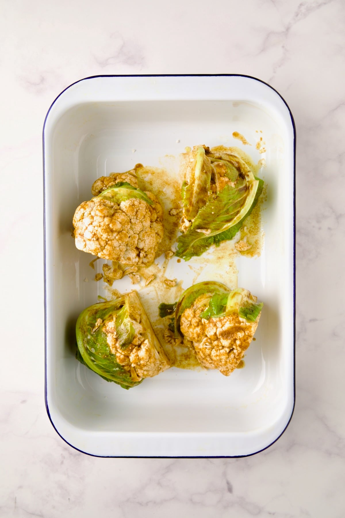 Four wedges of cauliflower in white roasting pan seasoned with curry powder before being roasted.